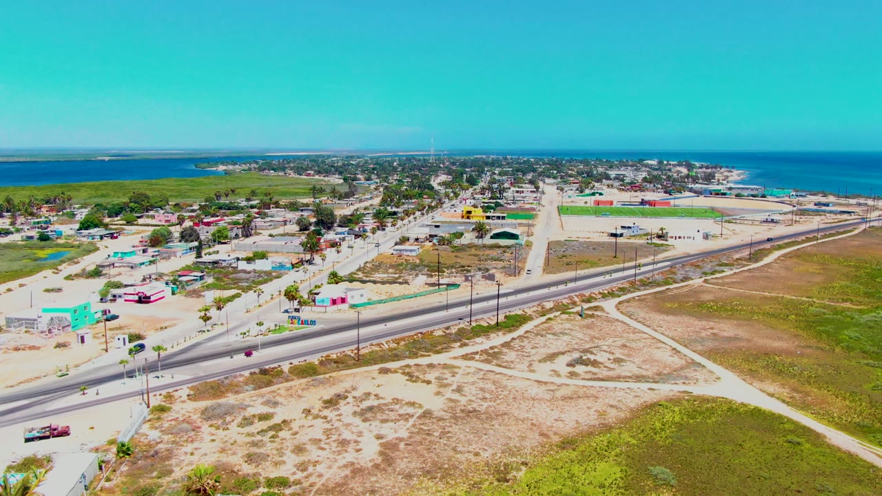 Wide drone frame in San Carlos, Baja California Sur, looking across scrubby lots and footpaths toward a highway with traffic, small town structures, green lowland, ocean, and no visible people.