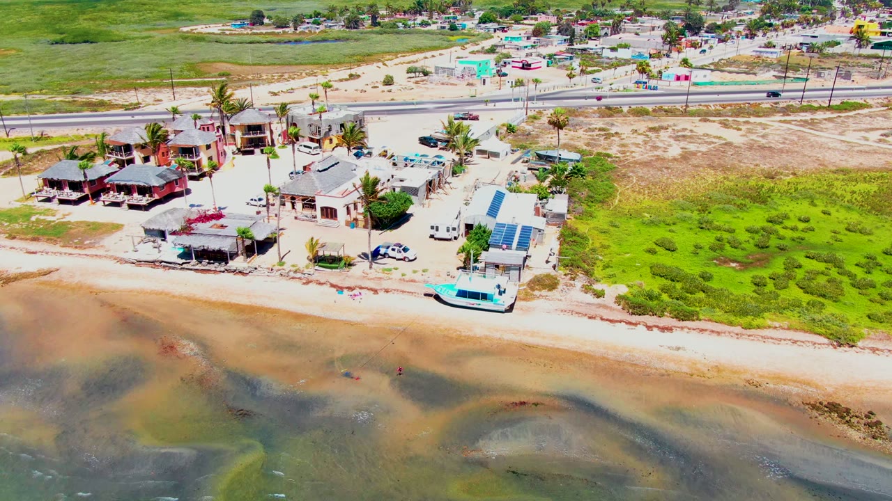 High aerial frame over San Carlos, Baja California Sur showing beachfront houses, a sandy strip, shallow patterned water, palms, parked vehicles, a road, and no visible people.