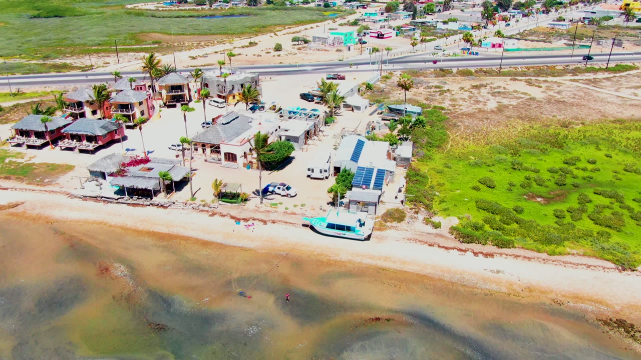 Drone view of beach houses, a white-roofed building, palms, road, sand, and shallow water.