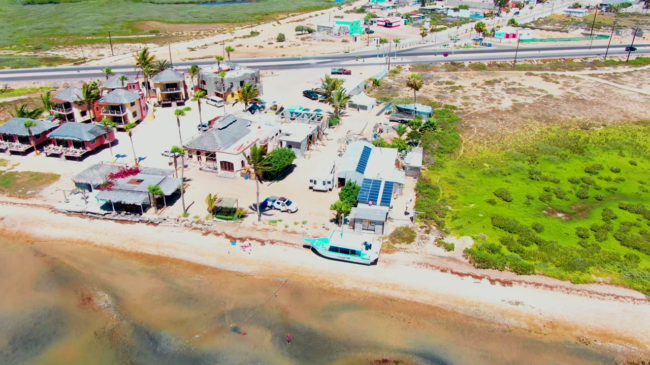 San Carlos, Baja California Sur aerial frame showing coastal homes centered near the beach, a road behind them, green open ground to the right, shallow water below, and no visible people.