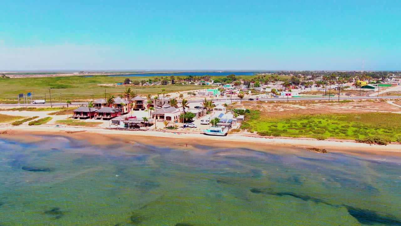Aerial coastline with beach houses, road traffic, grassy land, and shallow water.