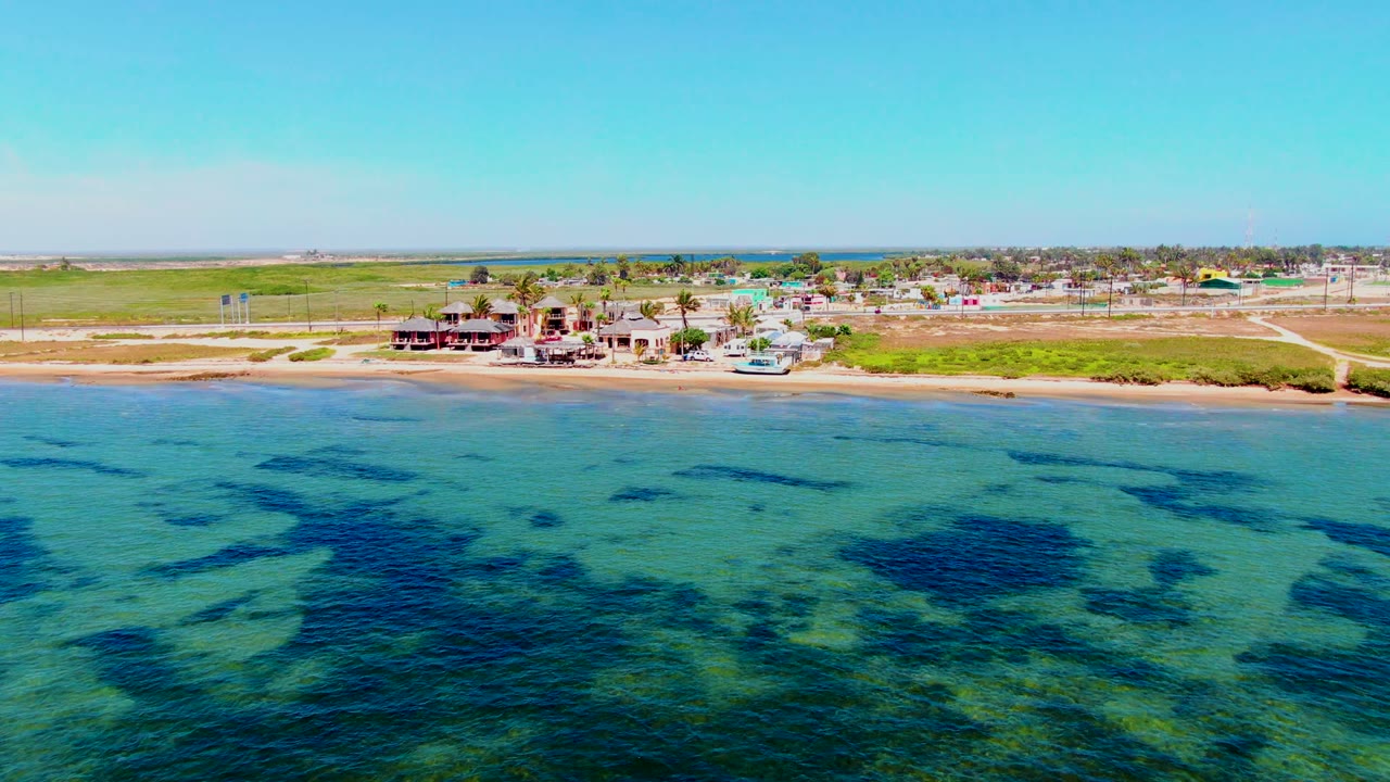 Opening aerial frame over San Carlos, Baja California Sur with clear turquoise shallows, dark underwater patches, a sandy beach, low coastal buildings, roads, grass, and no visible people.