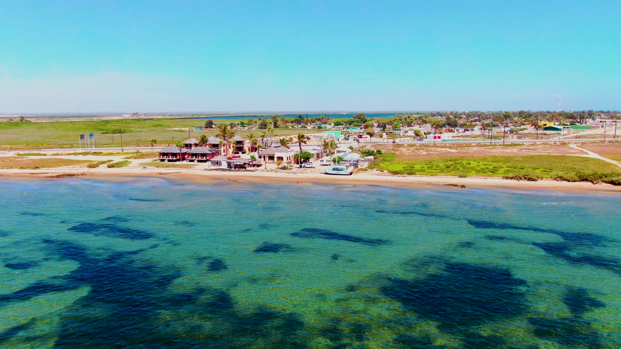 Mid-clip aerial frame over San Carlos, Baja California Sur with patterned shallow ocean in the foreground, sand and green grass along the coast, low buildings, roads, and no visible people.