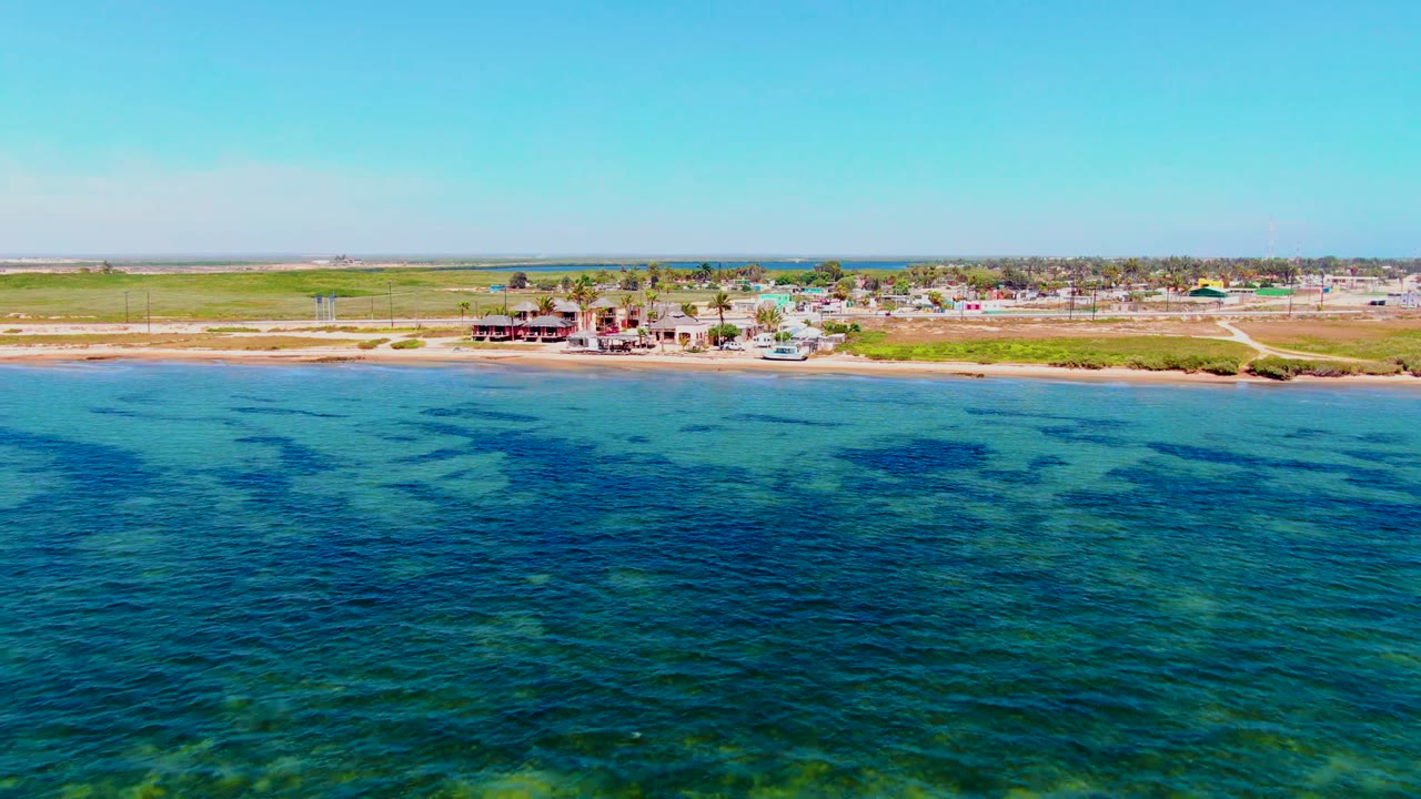 Opening aerial frame over San Carlos, Baja California Sur, with turquoise water in the foreground, a sandy shore and low buildings beyond it, and no visible people.