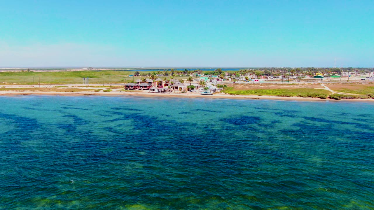 Wide aerial shoreline view with blue-green water, beach, and small coastal structures.