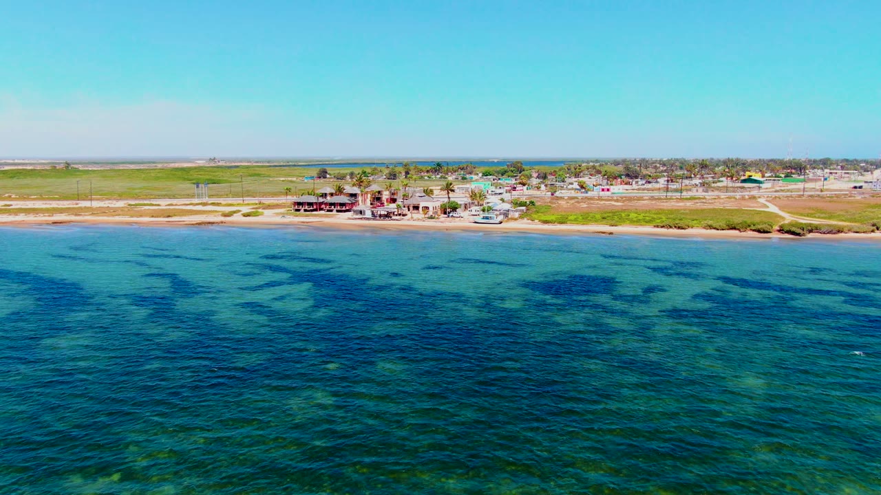 Aerial view in San Carlos, Baja California Sur, with clear turquoise water leading to a pale beach, scattered shore buildings, flat inland ground, and no visible people.