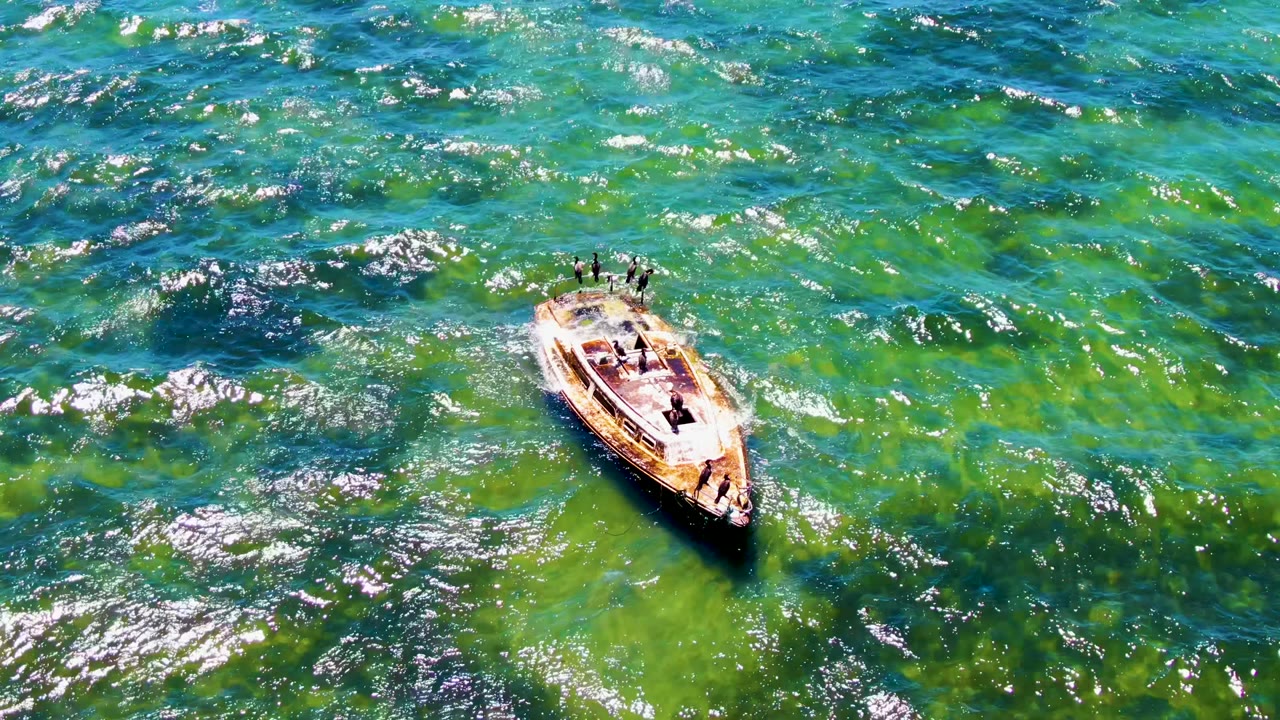 Aerial frame of a small motorboat crossing turquoise and green water in San Carlos, Baja California Sur, with people visible aboard and sun glitter on nearby waves.