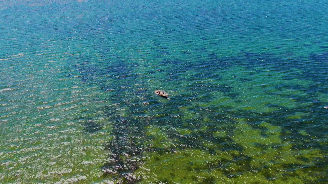Aerial view in San Carlos, Baja California Sur of a small boat near the left side of clear blue-green water with dark underwater rocks or coral patches and no visible people.