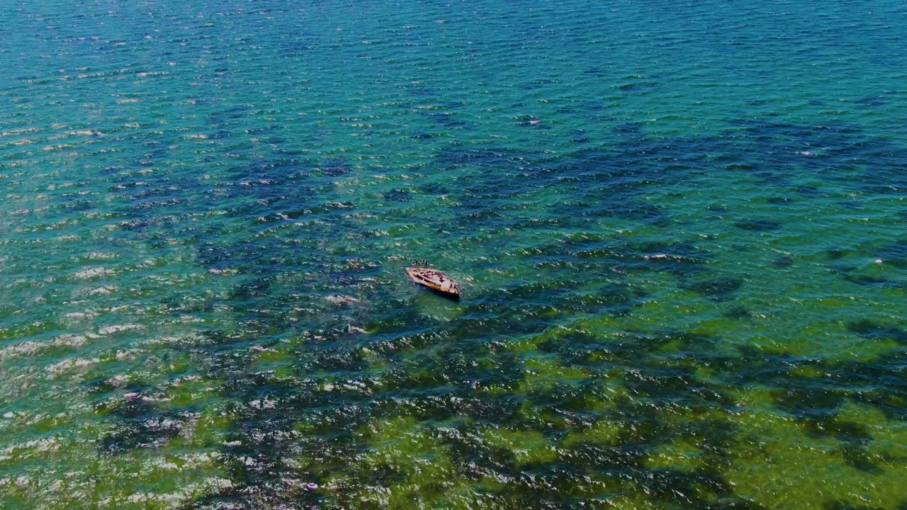 Aerial view in San Carlos, Baja California Sur with the boat near the upper right portion of the frame, floating above blue-green water and irregular dark underwater formations with no visible people.