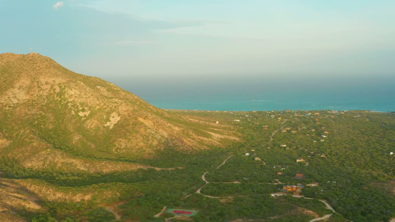 At 0:00, a wide aerial view over Cabo Pulmo shows a sunlit mountain on the left, green valley roads below, the distant sea on the horizon, and no visible people.