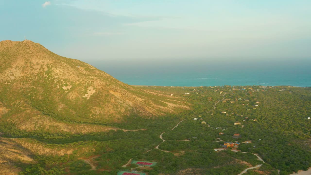 At 0:02, the aerial frame shows the mountain lit by low sun, a darker band of shadow across the lower hillside, rural roads in the valley, and the sea beyond, with no visible people.