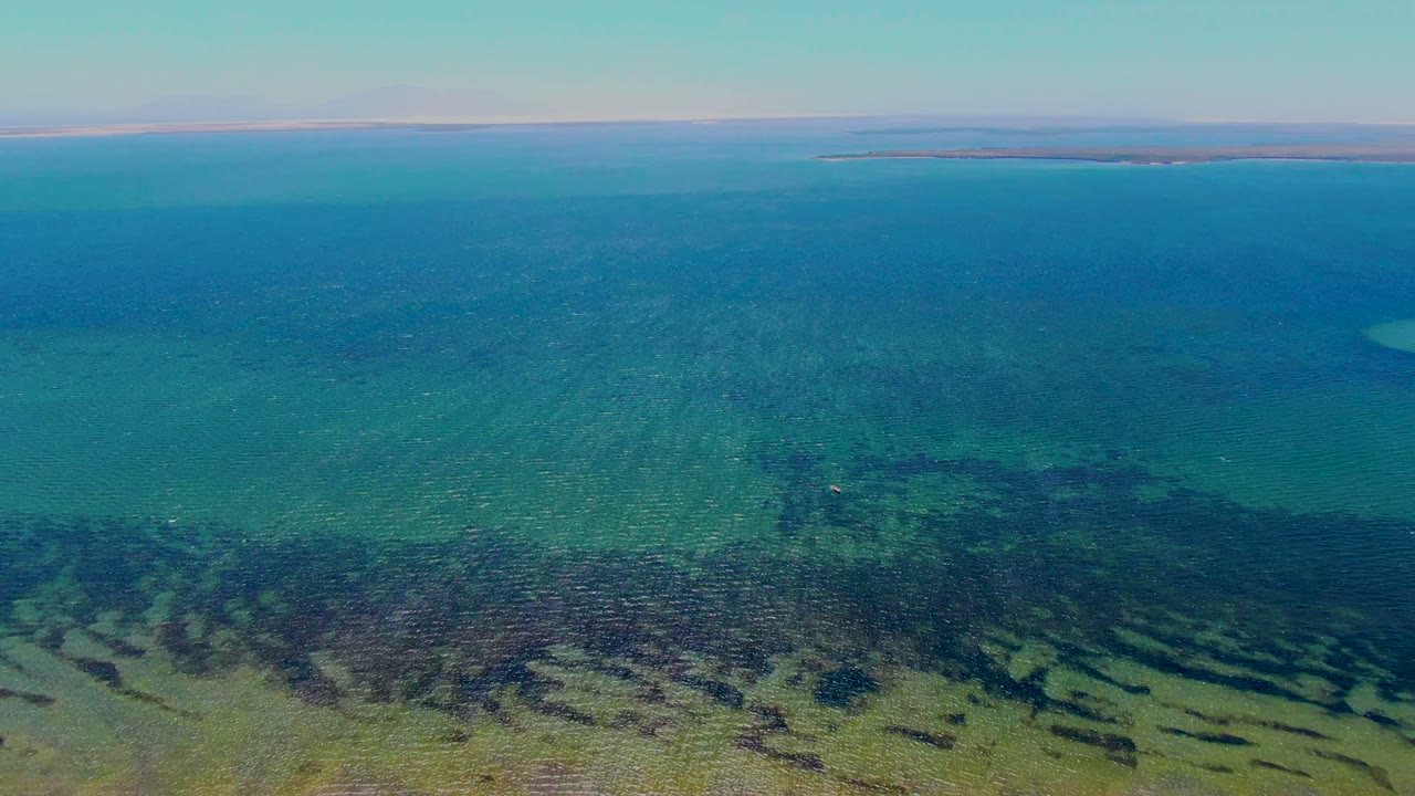 At 0:00, an aerial view over San Carlos, Baja California Sur shows turquoise shallow water, dark seabed texture, and a low island line on the horizon with no visible people.