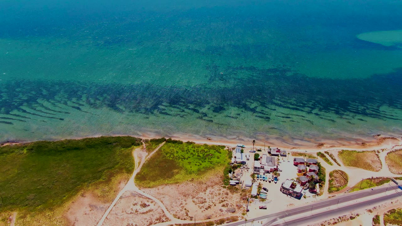 Aerial frame over San Carlos, Baja California Sur showing turquoise ocean above a sandy shoreline, green dunes, a road, small beachside buildings, and no visible people.