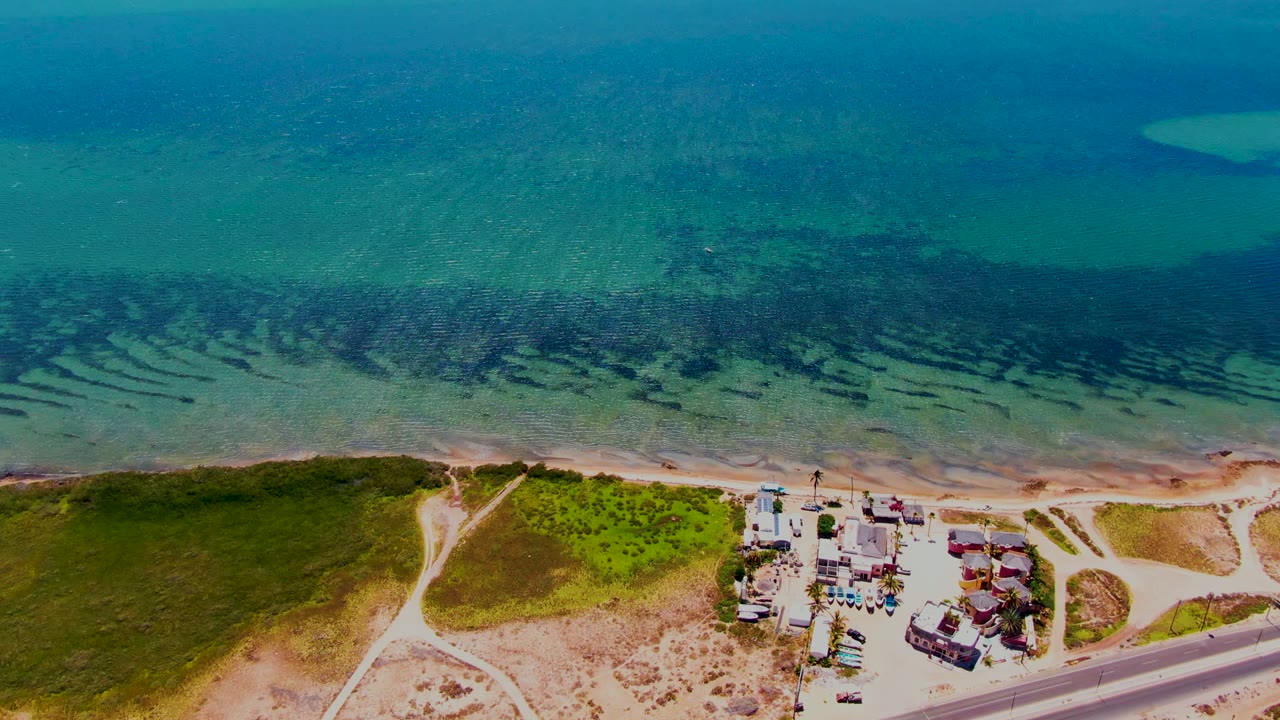 Turquoise coastal water above dunes, sandy shore, road, and clustered buildings.