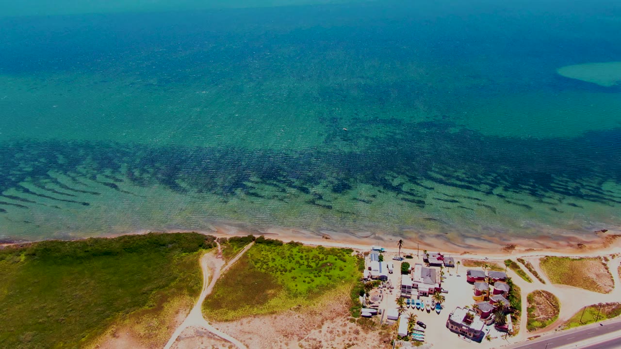 San Carlos aerial frame showing clear coastal water with dark underwater bands, a pale sandy shore, green dunes, roadside buildings near the bottom right, and no visible people.