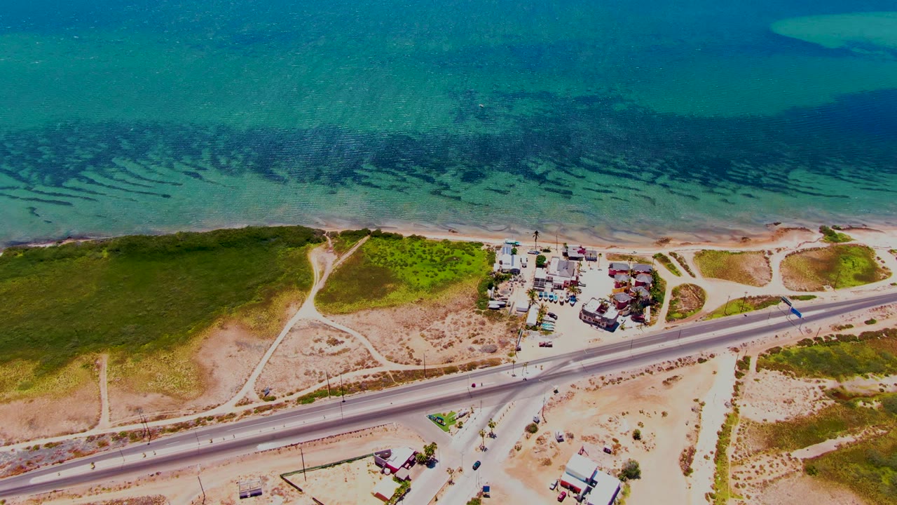 Aerial frame over San Carlos, Baja California Sur showing a coastal highway, sandy tracks, green scrub, a small building cluster, clear blue ocean, and no visible people.