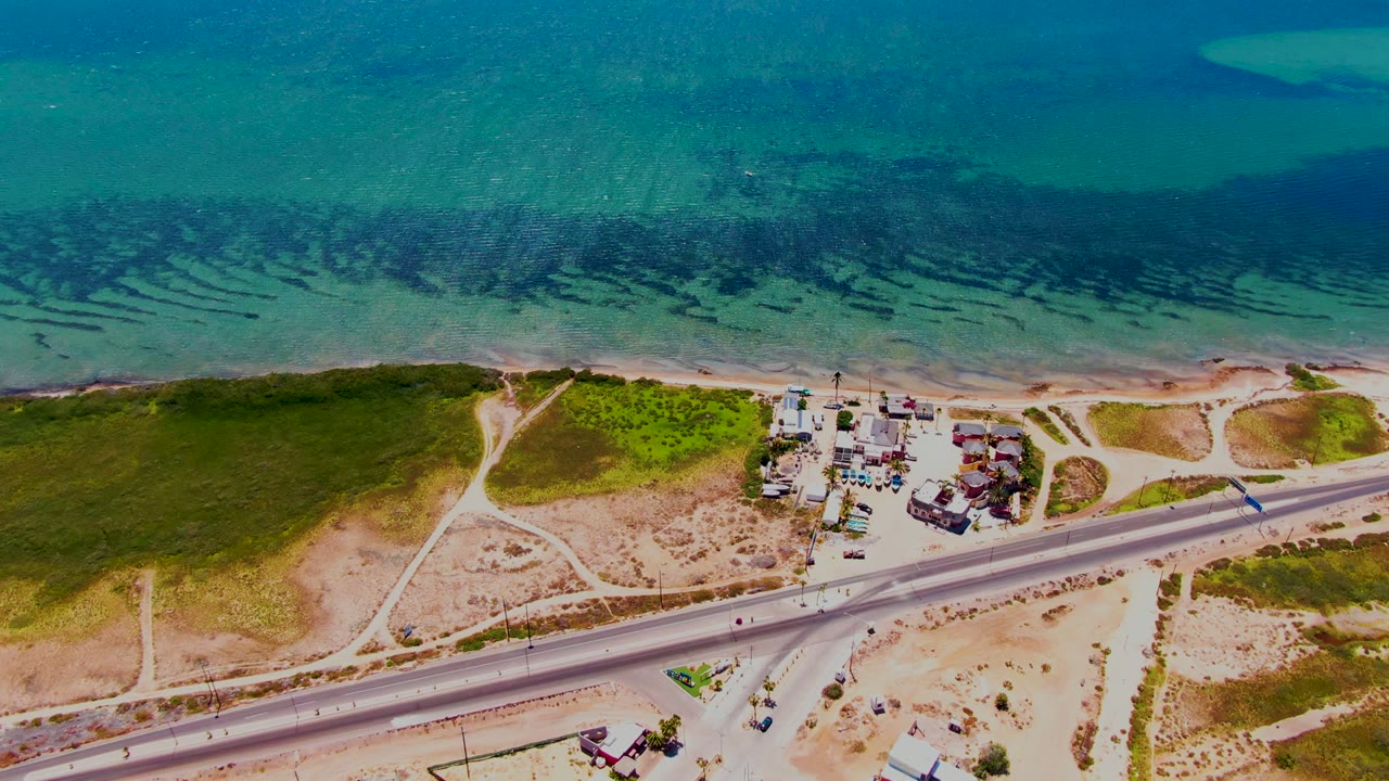 Overhead coastal road with vehicles, scrubland, buildings, and ocean water.
