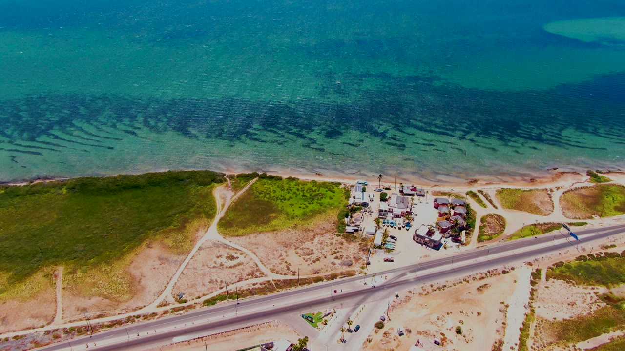 San Carlos, Baja California Sur aerial frame showing a highway through sandy coastal terrain, branching dirt paths, green patches, a compact settlement area, blue ocean water, and no visible people.