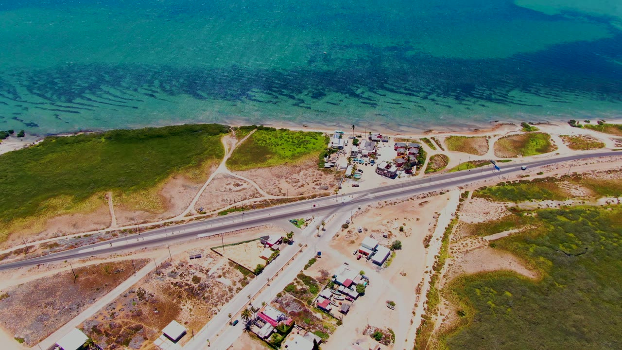 Opening aerial frame over San Carlos, Baja California Sur, showing turquoise ocean, a sandy shoreline, a paved highway, scattered buildings, dirt tracks, and no visible people.