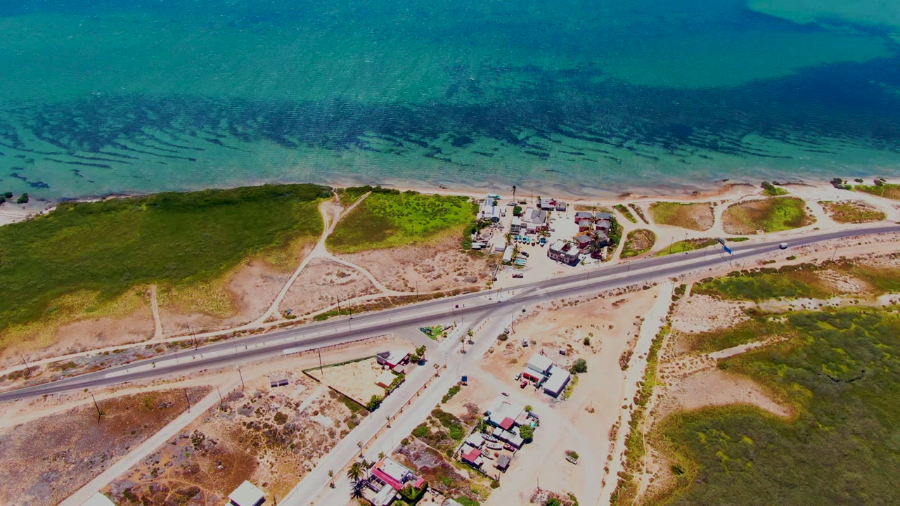 Overhead coastal road with vehicles, scrubland, and clear blue water.
