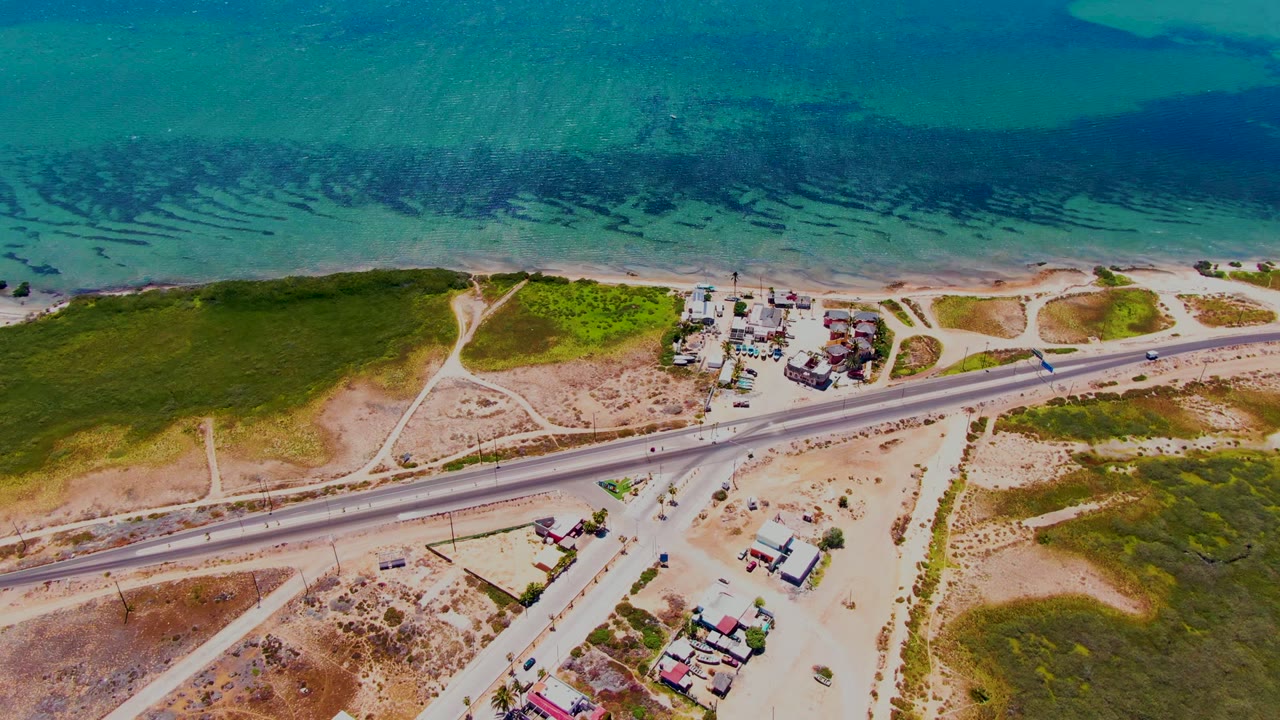 Aerial view in San Carlos, Baja California Sur, showing the coastal highway angled across sandy ground, vehicles in both lanes, clustered roadside buildings, blue ocean, and no visible people.