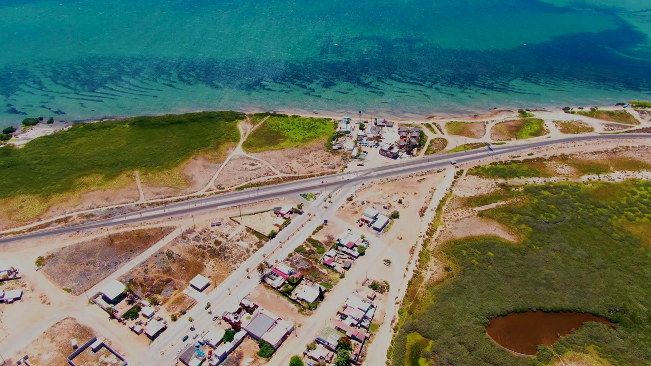 Aerial frame over San Carlos, Baja California Sur showing turquoise ocean, a narrow sandy shoreline, a coastal highway, green scrubland, scattered homes, and no visible people.