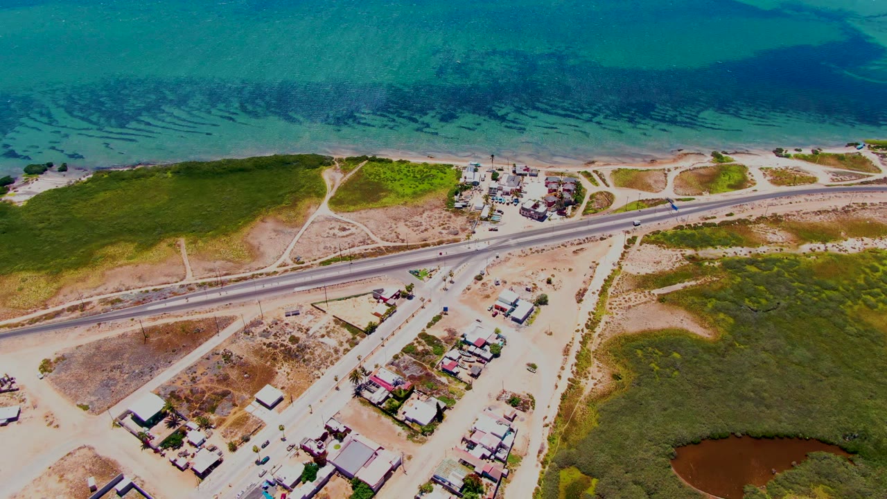 Drone view of a coastal highway beside clear water, scrubland, dirt roads, and houses.