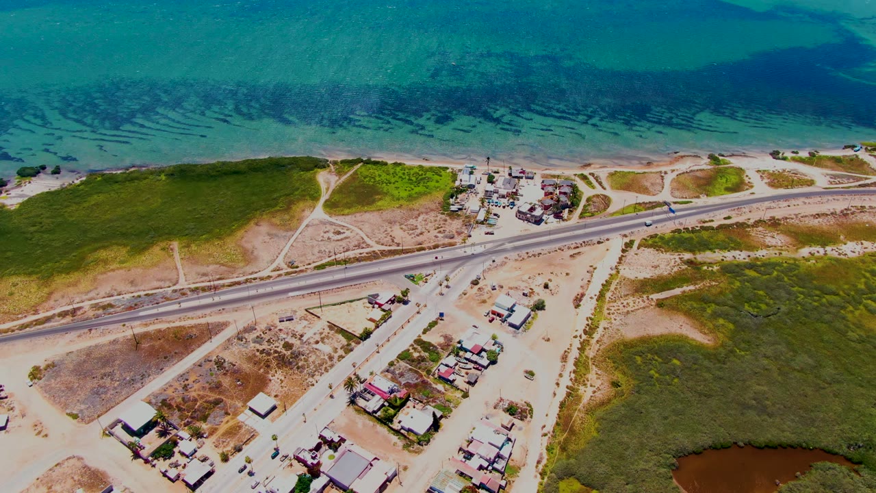Aerial frame over San Carlos, Baja California Sur showing blue-green coastal water, a sandy beach strip, the highway, open scrubland, clustered homes, and no visible people.
