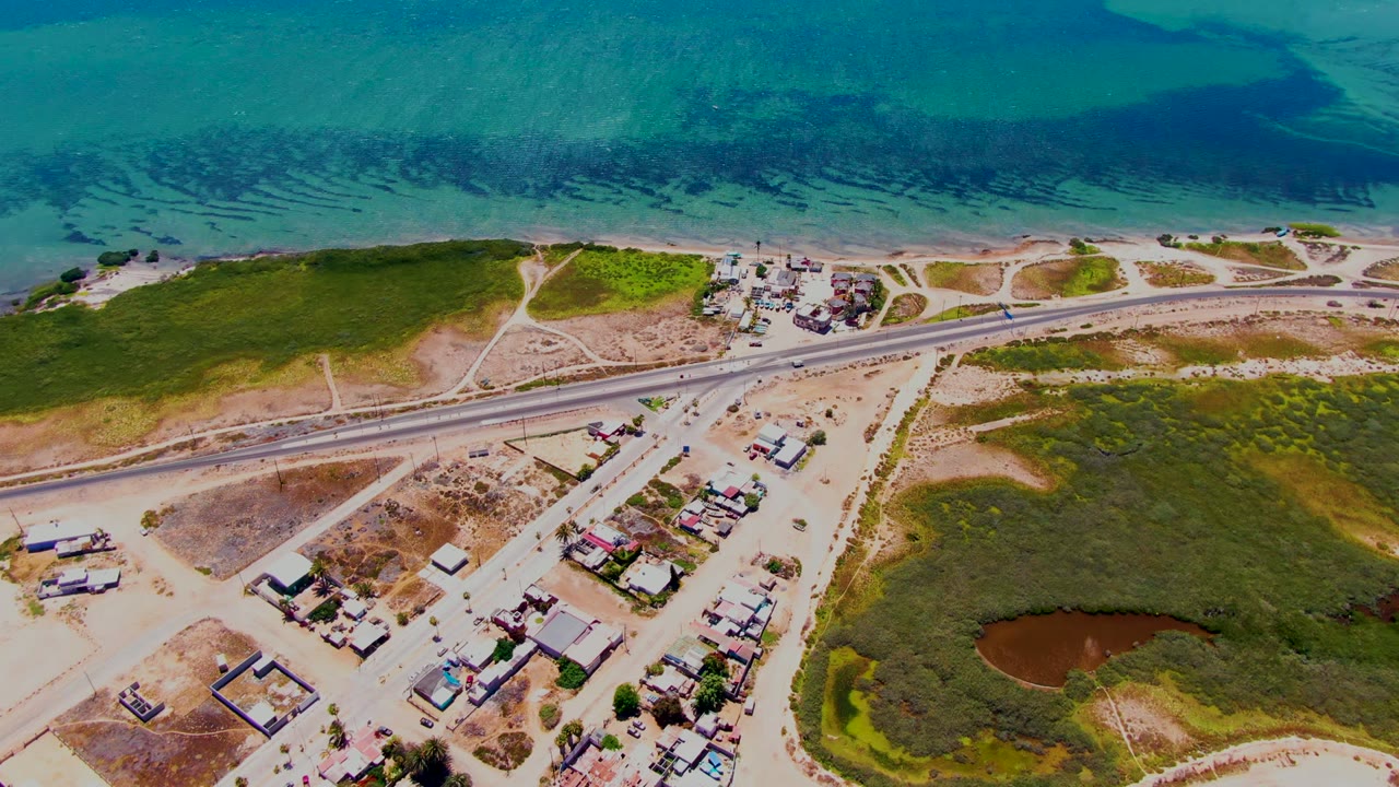 Opening aerial frame over San Carlos, Baja California Sur, showing blue ocean, sandy shore, a parallel road, scattered buildings, green terrain, and a small pond area with no visible people.