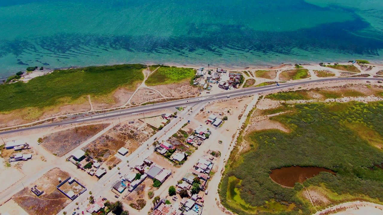 Aerial view over San Carlos, Baja California Sur showing clear blue water, a sandy strip, the coastal road with small vehicles, clustered buildings, green ground cover, and a brown pond with no visible people.