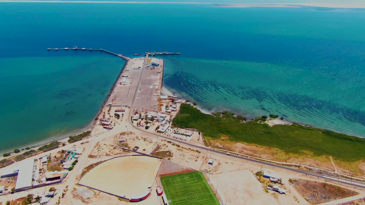 Aerial frame over San Carlos, Baja California Sur showing a long dock reaching into blue-green water, pale shoreline land, a curved-roof building, a green field, and no visible people.
