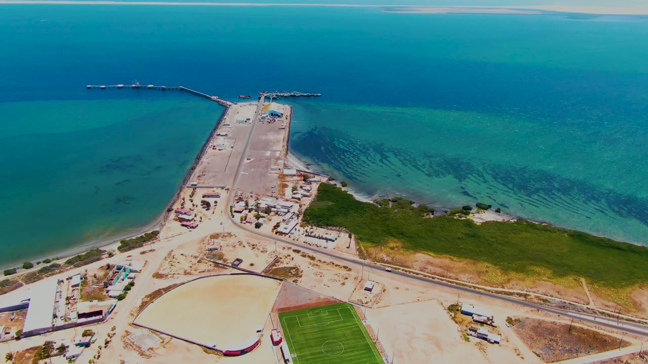 Elevated coastal view with the dock centered, shallow water on both sides, and land activity below.