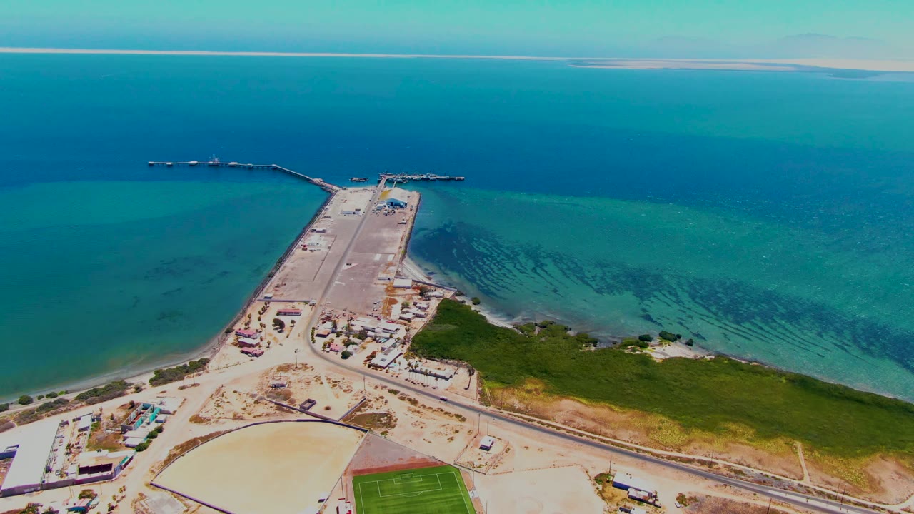 Aerial frame over San Carlos, Baja California Sur showing a causeway and pier extending into blue-green ocean, with sandy shore and a green field below, no visible people.