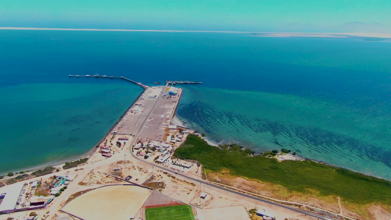San Carlos aerial frame showing the pier head near the top of the causeway, green shoreline vegetation at lower right, sandy road areas, blue water, and no visible people.