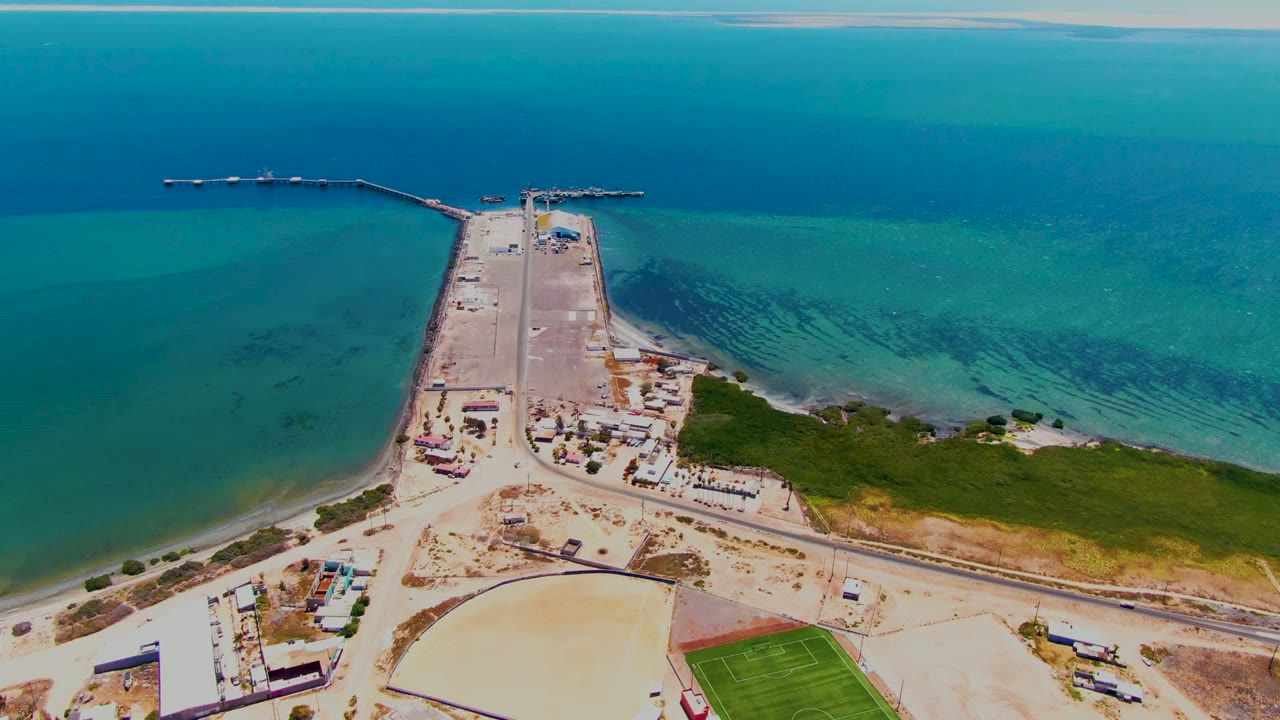Early aerial frame over San Carlos, Baja California Sur showing the pier centered into blue-green ocean, shoreline roads below, a green field at lower right, and no visible people.