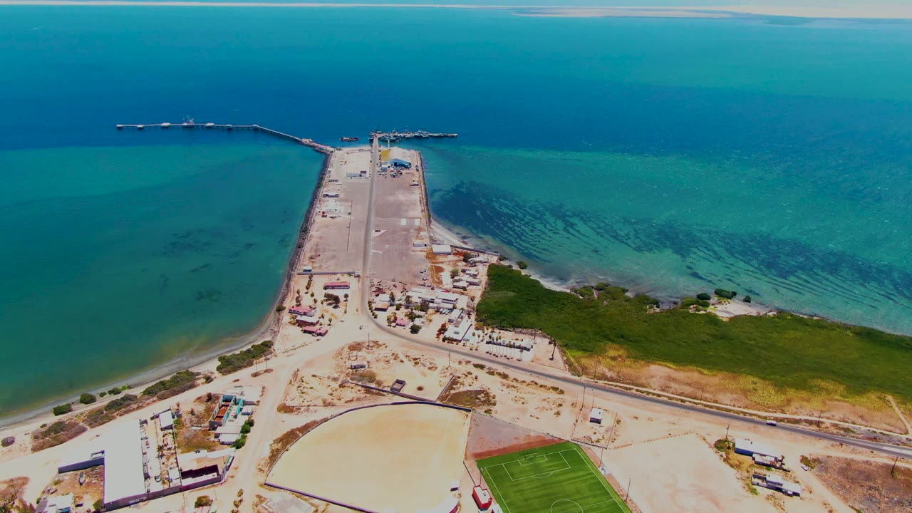 Drone view of the coastal pier with blue water, shore road, buildings, and a green field.