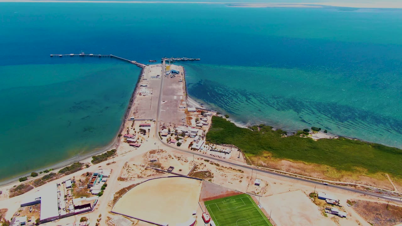 San Carlos aerial frame showing the pier projecting into darker blue ocean, rippled shallow water near shore, sandy lots and a green field below, with no visible people.