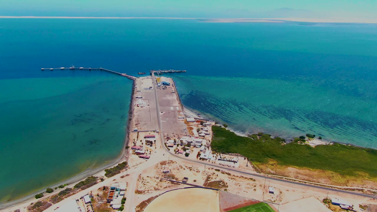 High aerial frame over San Carlos, Baja California Sur showing a pale runway or paved strip extending into turquoise ocean, with sandy land, jetties, and no visible people.