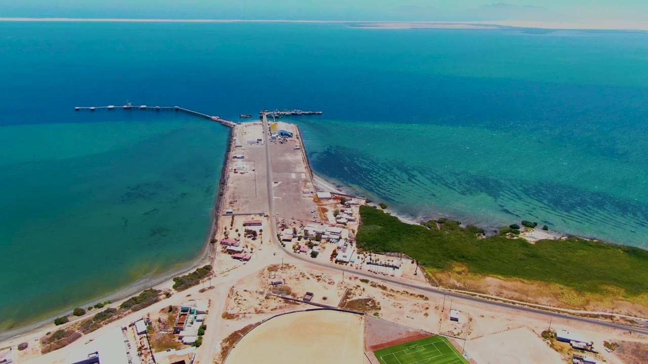 High drone-style frame over San Carlos, Baja California Sur showing the runway leading to the ocean, a curved shoreline, sandy lots, a green field at the lower edge, and no visible people.