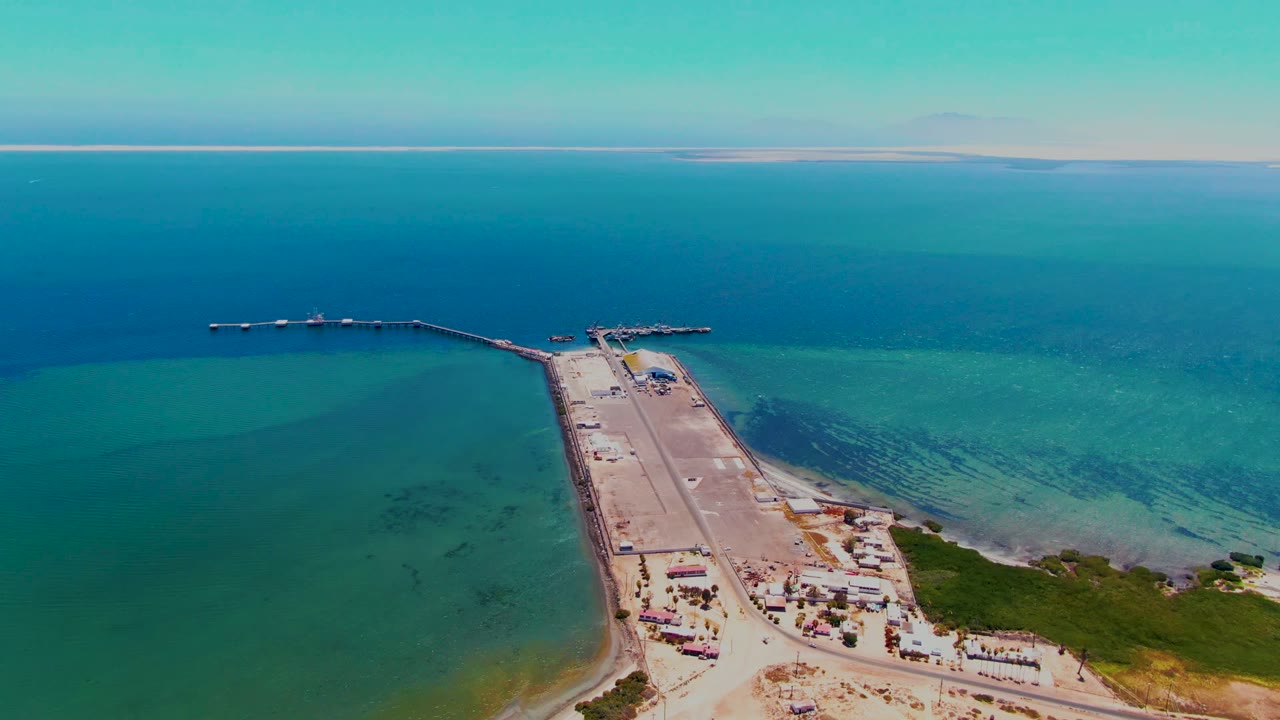 Aerial frame from San Carlos, Baja California Sur showing a long pier reaching into turquoise and deep blue ocean water, with sandy shore, road, small structures, and no visible people.