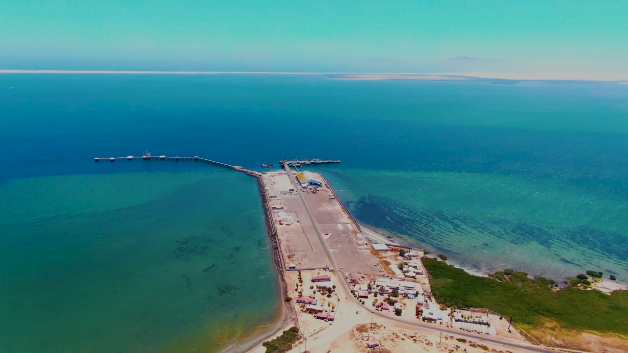 Drone-style frame from San Carlos, Baja California Sur showing the pier extending from the lower right shore into blue water, with a road, small coastal structures, scrub vegetation, and no visible people.