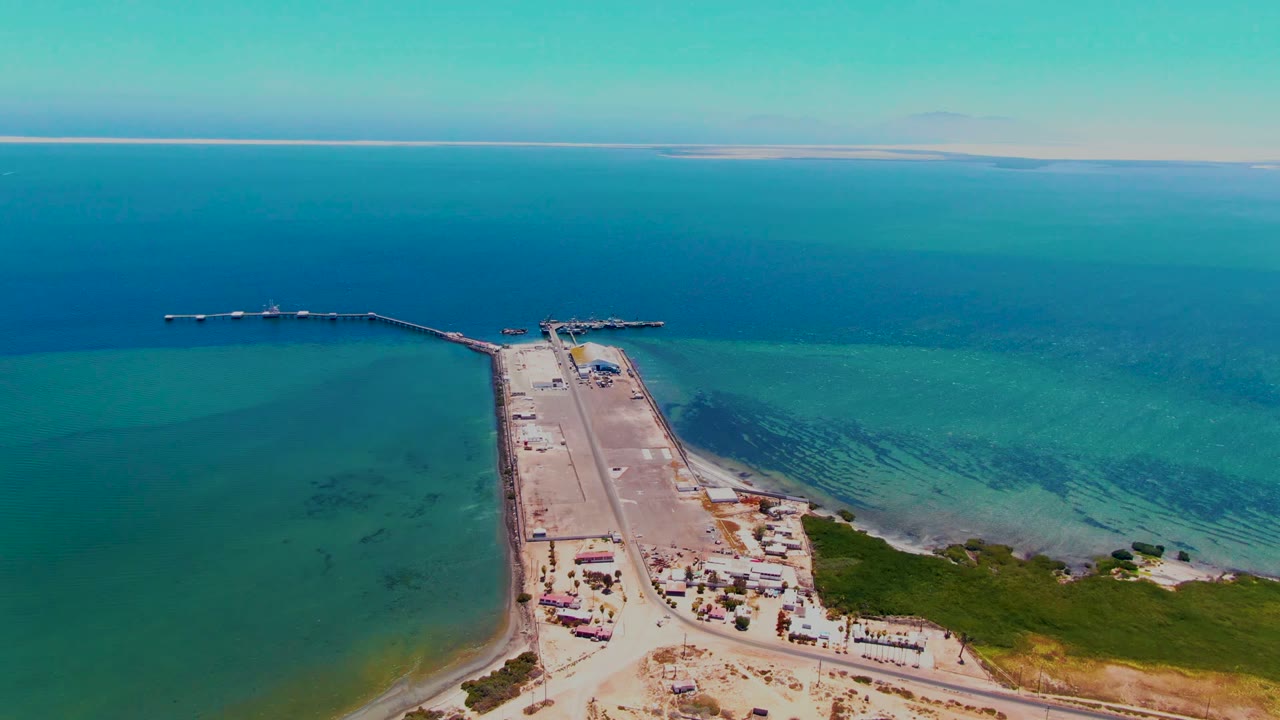 Aerial frame over San Carlos, Baja California Sur, showing a concrete dock and jetty reaching into turquoise ocean, pale shore in the foreground, and no visible people.