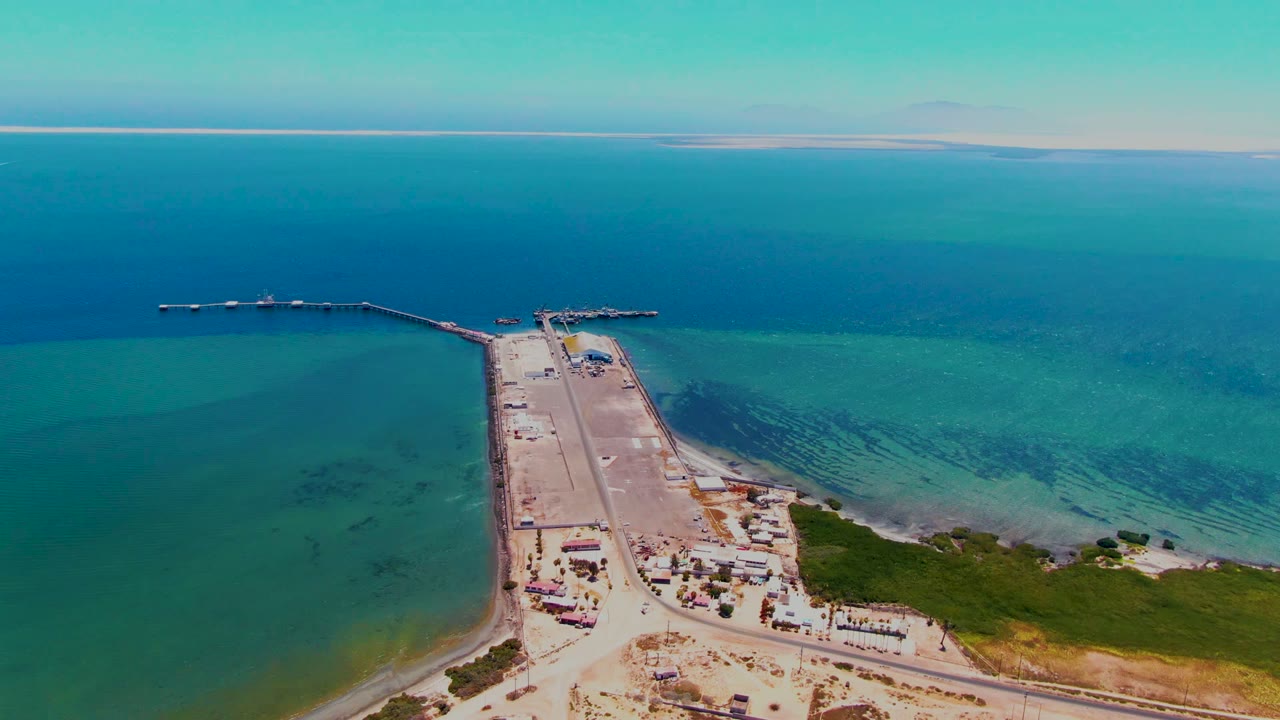 A high drone view in San Carlos, Baja California Sur, centers the dock approach with blue-green water on both sides, small shore structures below, and no visible people.