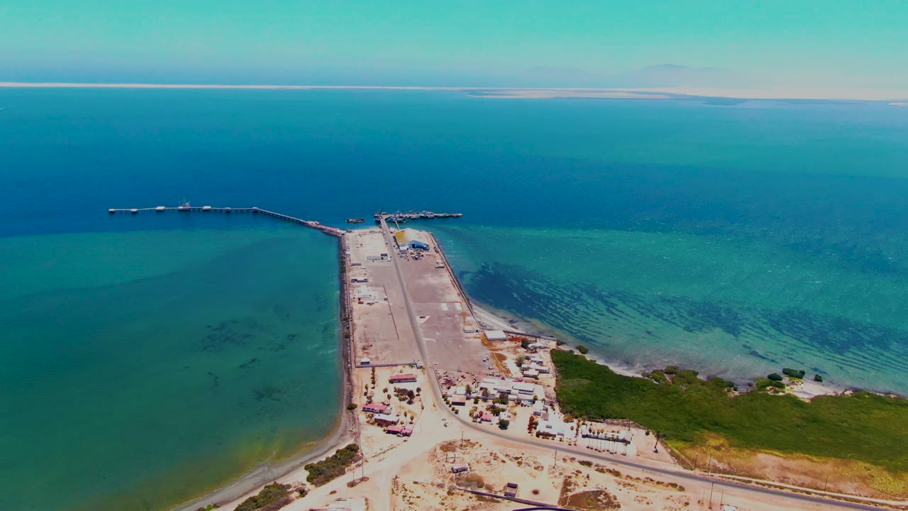 The San Carlos aerial frame shows the long dock leading to small jetty arms, darker underwater patches in the turquoise sea, shoreline construction areas, and no visible people.