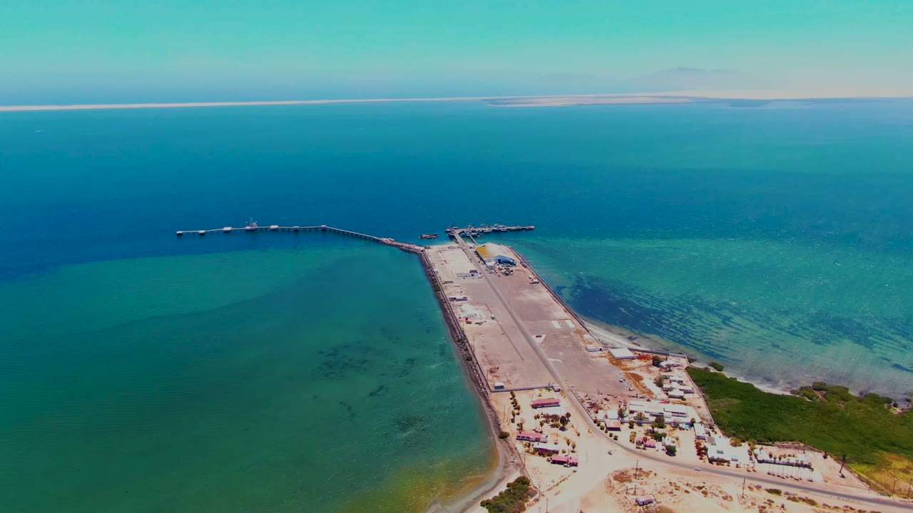 High aerial view over San Carlos, Baja California Sur, with a long dock extending from sandy shore into turquoise and blue ocean, no visible people.