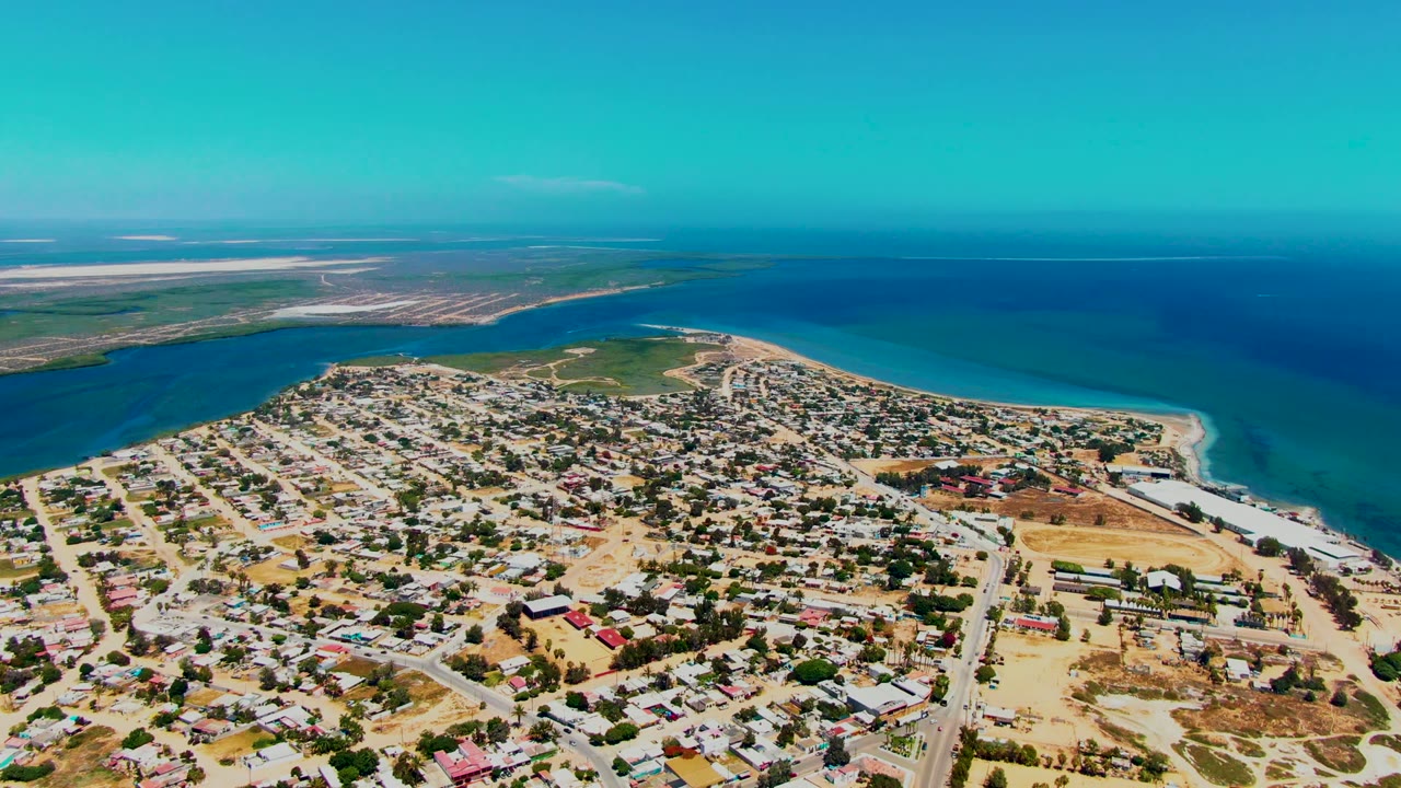 High aerial view over San Carlos, Baja California Sur, showing residential blocks, roads, sandy beach, blue-green coastal water, darker ocean beyond, and no visible people.
