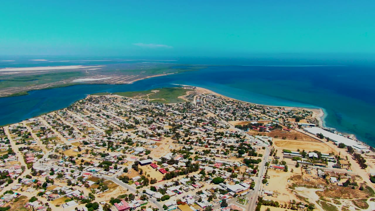 Wide drone view of houses, roads, beach, inlet, and calm ocean water.