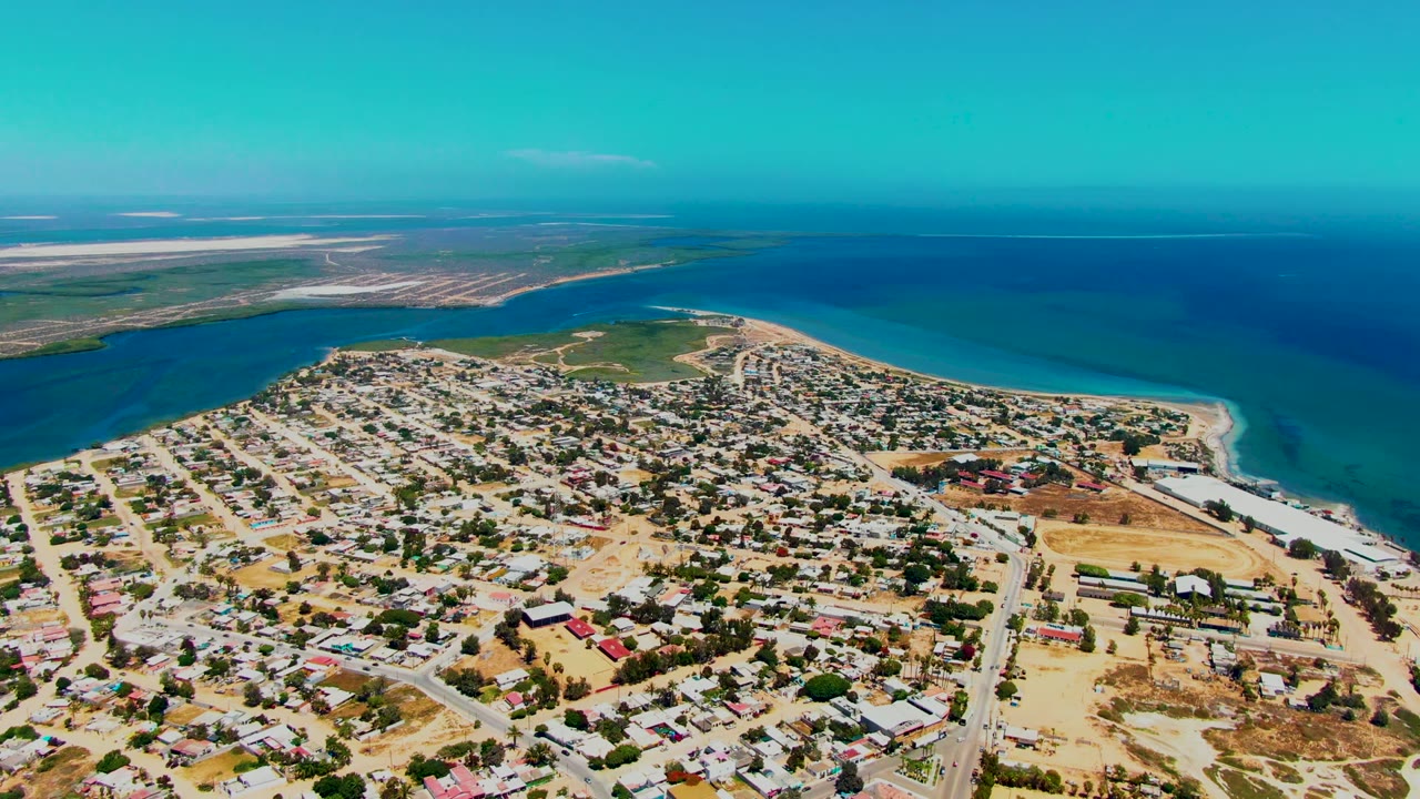 Drone view above San Carlos, Baja California Sur, looking across low residential buildings toward a sandy point, turquoise nearshore water, darker sea, flat coastal land, and no visible people.