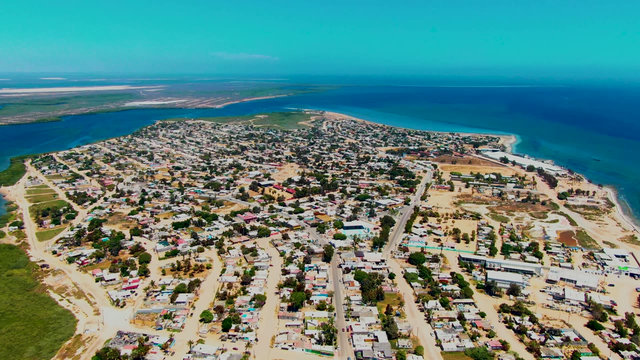 Wide drone view over San Carlos, Baja California Sur, showing residential blocks, sandy roads, a curved beach, turquoise water, darker ocean, and no visible people.