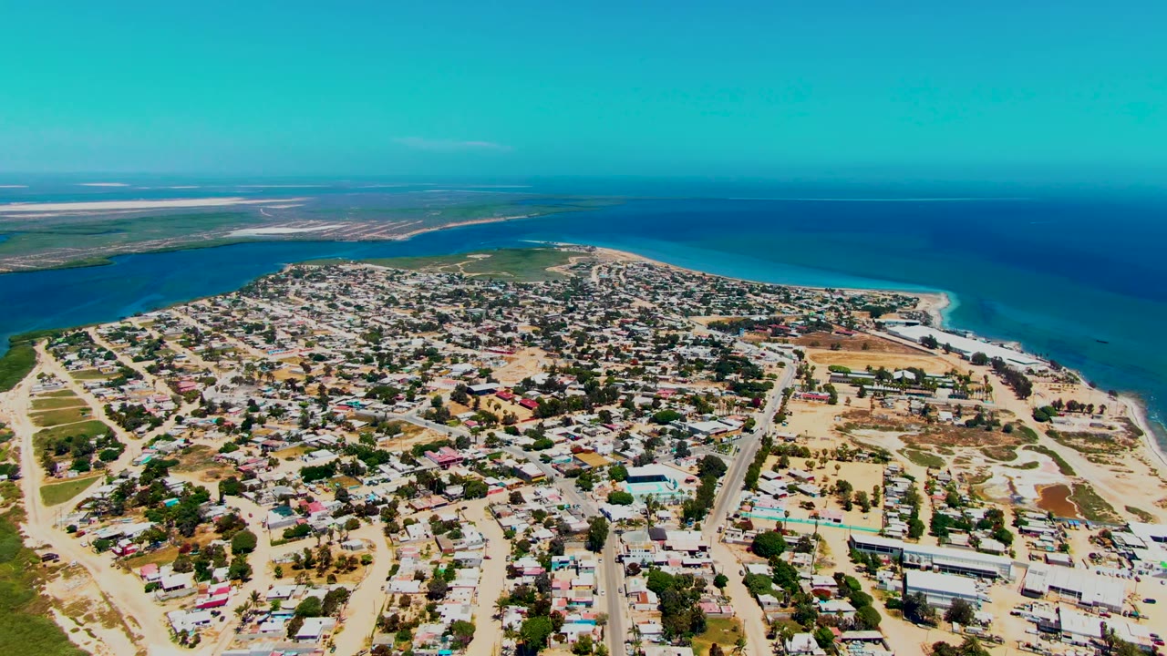San Carlos appears from a slightly shifted drone angle with low houses, pale streets, open sandy parcels, bright beach edge, blue ocean, and no visible people.